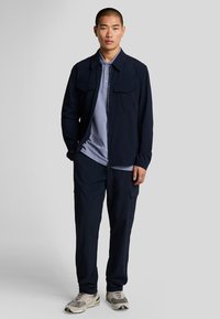 Giacca zip in navy con due tasche frontali, abbinata a una polo viola chiaro e pantaloni navy. Indossata con sneakers beige e grigie.