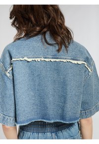 Giacca di jeans tagliata di un blu medio con orlo sfrangiato e dettagli usurati sulle spalle. Include maniche oversize e cuciture.