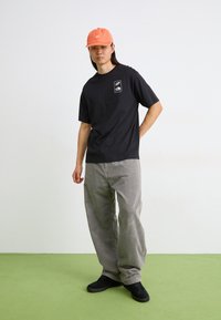 Black short-sleeve t-shirt with a logo patch, paired with gray corduroy pants. The model wears a coral cap and black shoes. Green floor below.