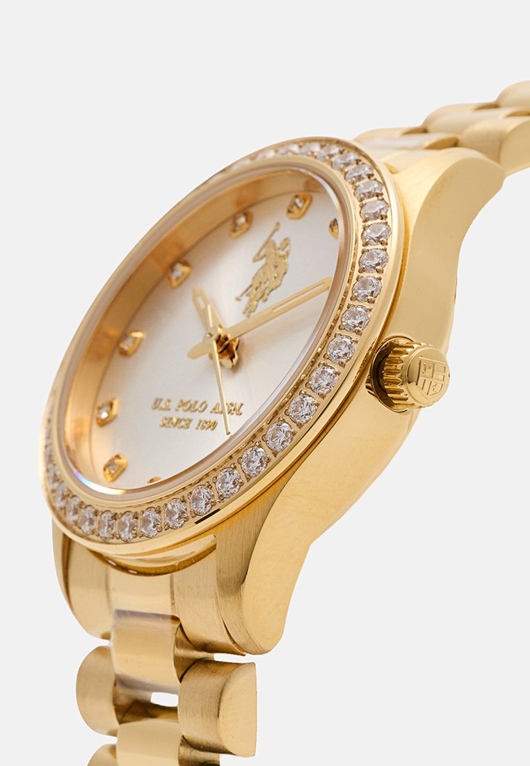 Montre dorée avec un cadran rond, un cadran blanc, orné de cristaux transparents et d'index en or. Elle est dotée d'un bracelet à maillons et d'une couronne.