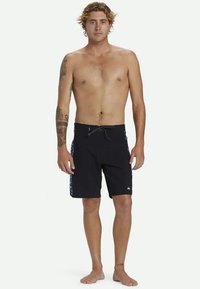 Homem sem camisola com tatuagens, a pé descalço, vestindo calções de banho pretos com padrões laterais e cordão na cintura, contra um fundo branco.