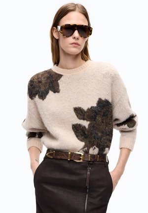 Donna con occhiali da sole oversize tartarugati, maglione beige con grandi motivi floreali scuri, gonna in pelle marrone e cintura con fibbia dorata.