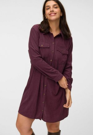 Femme portant une robe-chemise à manches longues boutonnée violet foncé avec des poches poitrine, ajustant une manche, souriant sur un fond clair.