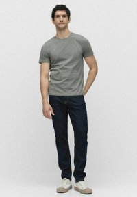 Grå kortærmet t-shirt, mørke jeans og lyse sneakers. Enkel design, tætsiddende form og glat tekstur. Stående pose mod en neutral baggrund.