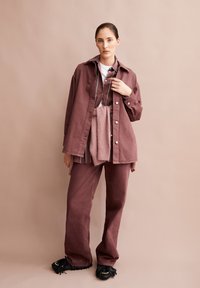 Frau in lockerer mauvefarbener Jacke und Hose über gestreiftem Hemd, mit einem Pullover um die Taille gebunden, vor beigem Hintergrund stehend.