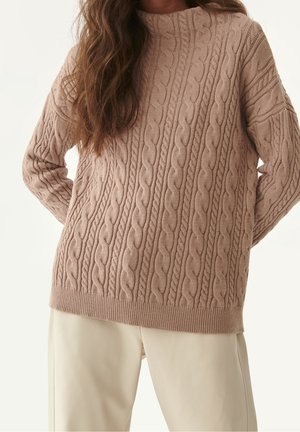 Woman wearing a beige cable-knit sweater with long sleeves and cream-colored pants, arms bent behind her back.