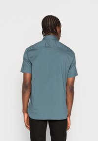 Camicia a maniche corte in tessuto teal con una superficie liscia, caratterizzata da un colletto classico e orlo dritto. Senza pattern o dettagli visibili.