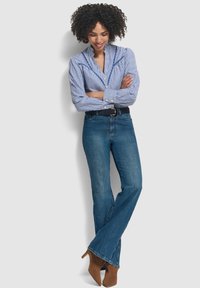 Camicia a righe blu con taglio aderente, abbinata a jeans blu a zampa d'elefante e stivali con tacco marroni, il tutto su uno sfondo neutro.