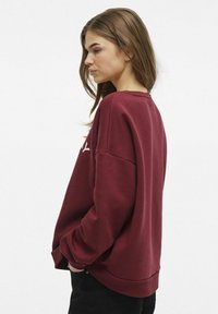 Felpa bordeaux con vestibilità morbida, polsini e orlo a costine. Presenta spalline scese e un piccolo logo bianco sulla parte anteriore. Tessuto morbido.