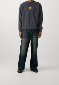 Person med en urtvättad svart sweatshirt med en gul sol-logga och vida urtvättade blå jeans, som står mot en neutral ljus bakgrund.