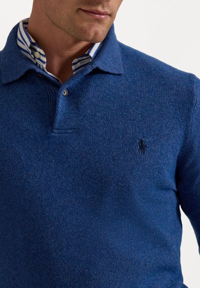 Blå strikket polo sweater med en struktureret overflade, der har en krave, to knapper og en broderet logo på venstre bryst.