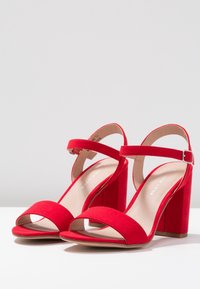 Red suede high-heeled sandals with an ankle strap, open toe, and square block heel. Smooth inner lining and stitched detailing.