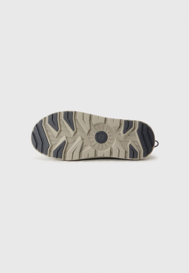 Semelle de chaussure en beige et bleu foncé, présentant un motif texturé et robuste avec des rainures pour une meilleure adhérence et un logo circulaire au centre.