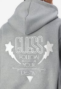 Pilkas su kapučiu džemperis su baltais spausdintais žodžiais: "GUESS FOLLOW YOUR DESTINY," su žvaigždžių ir žaibo grafikomis. Minkšta tekstūra.