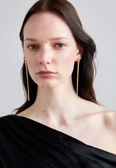 Pendientes largos de tono dorado, con un diseño minimalista y elegante, acabado suave, que complementan la blusa negra del modelo.