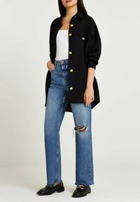 Svart texturerad skjortjacka med guldknappar, kombinerad med ljusblå högmidjade trasiga jeans och svarta skor. Enkel vit linne under.