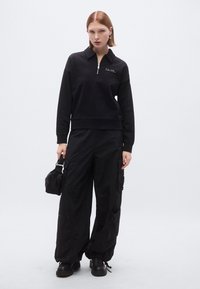 Felpa nera con zip e collo alto, con logo ricamato, abbinata a pantaloni cargo neri larghi. Presenta multiple tasche e una vestibilità comoda.