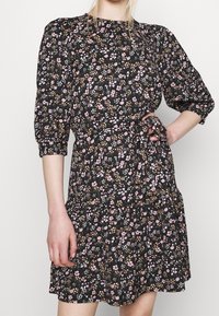 Vrouw in een zwart jurkje met een roze en beige bloemenpatroon, met opbollende driekwart mouwen en een strik in de taille, met één hand op de heup.