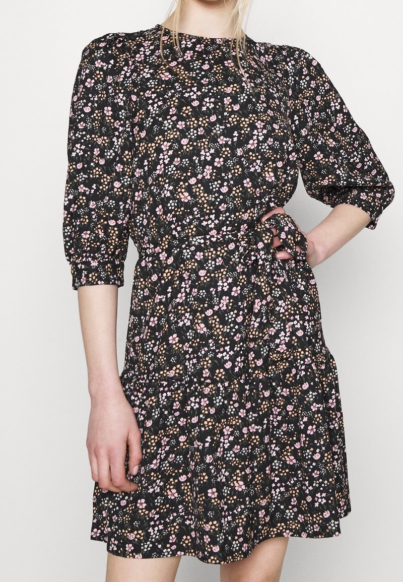 Vrouw in een zwart jurkje met een roze en beige bloemenpatroon, met opbollende driekwart mouwen en een strik in de taille, met één hand op de heup.