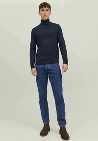Jovem em pé a usar um suéter de gola alta azul marinho, jeans azuis e botas castanhas de tornozelo contra um fundo claro e liso.