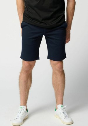 Muž oblečený v námornícky modrých šortkách, čiernom tričku a bielych teniskách so zelenými detailmi, stojaci pred jednoduchým svetlým pozadím.