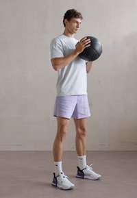 Mandens atletiske outfit består af en lysegrå T-shirt, lavendel shorts, hvide sokker og sort/hvide sneakers, mens han holder en sort basketball.