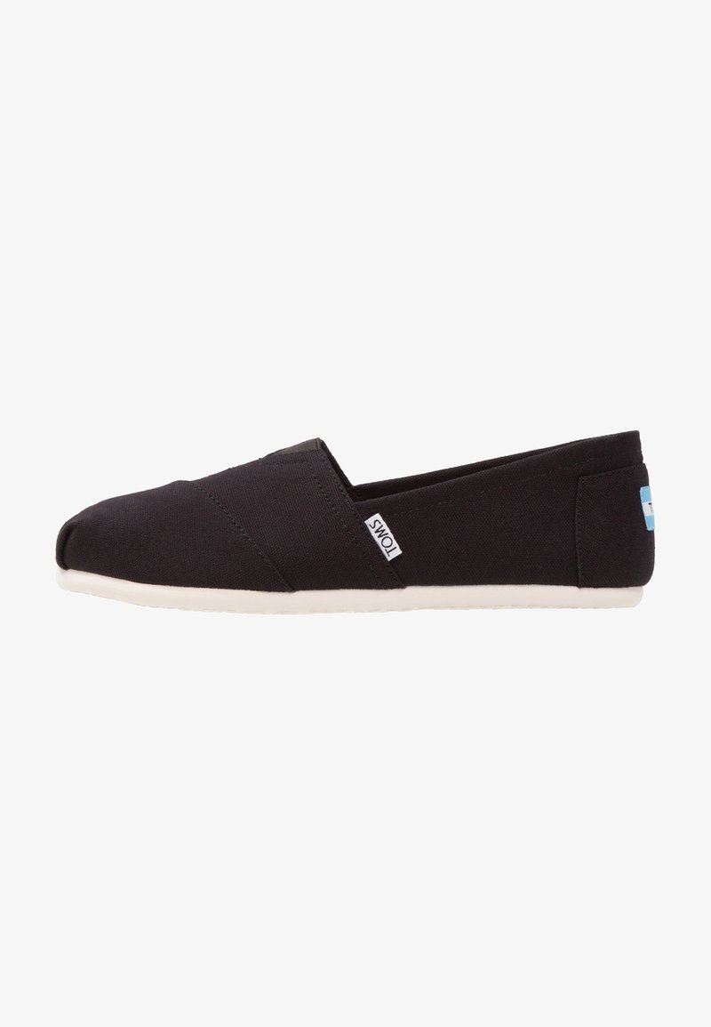Chaussure en toile noire sans lacets avec semelle blanche, avec une petite étiquette blanche « TOMS » sur le côté et un logo bleu au talon.