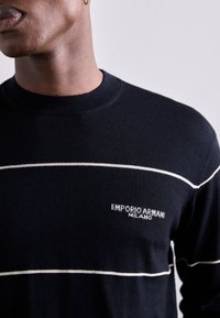 Maglione di lana blu scuro con strisce orizzontali bianche. Presenta un collo a giro e il logo ricamato "EMPORIO ARMANI MILANO" sul petto.