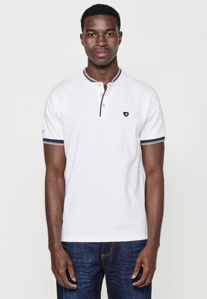 Polo bianco con colletto alto, patta con bottoni, dettagli a righe blu navy e rosse, abbinato a jeans in denim scuro.