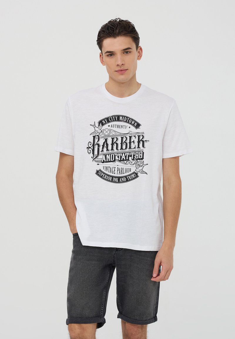 T-shirt bianca in cotone con una grafica nera di un barbiere vintage e un tatuaggio. Indossato con pantaloni corti di denim scuri, che mostrano un look casual.