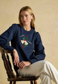 Jūras spēku kreklis ar pūkām piedurknēm un izšūtu svētku dizainu ar zvaniņiem un efeju. Ribotās piedurknes un apakša piešķir tekstūru.