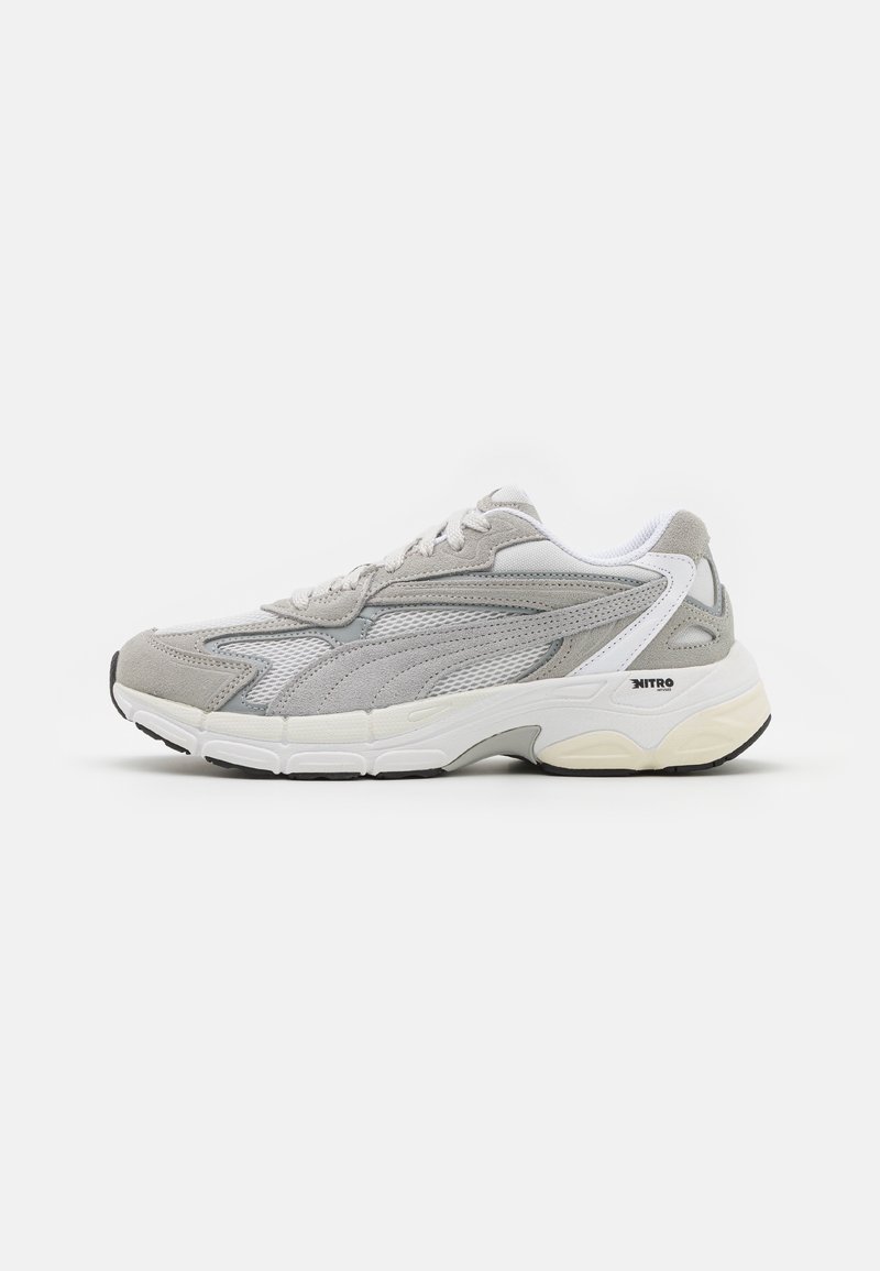Gray athletic sneaker with mesh and suede materials, featuring a thick white midsole, textured accents, and flat black outsole. Lace-up design.