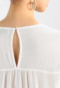 Blouse blanche texturée avec une fermeture à boutons au dos et un détail découpé. Présentant un tissu doux et léger ainsi qu'un ourlet évasé.