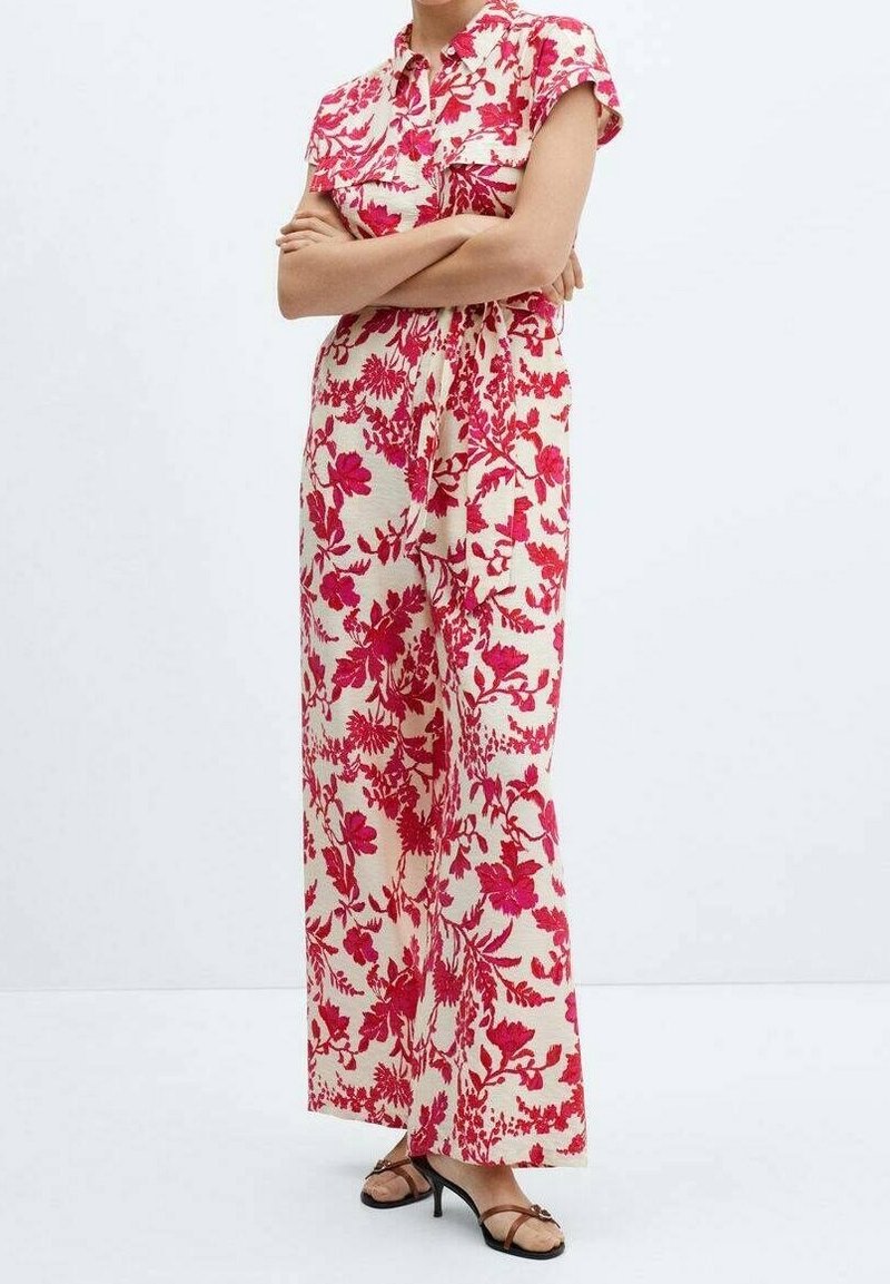 Femme portant une combinaison sans manches avec des motifs floraux rouges sur fond blanc et des sandales noires à talons, debout, bras croisés.