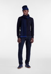 Mornarsko pleten pulover s teksturiranim vzorcem, ujemajoča šala, oprijete temne kavbojke in mornarski športni čevlji s svetlimi podplati, postavljeni na nevtralno ozadje.