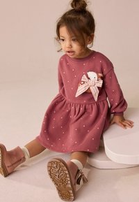 Roze jurk met stippen, lange mouwen, geplooide taille en een stoffen beer met een gestreepte strik. Gecombineerd met roze enkellaarsjes en witte sokken.