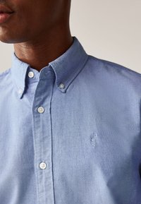 Hellblaues Hemd mit Knopfleiste und Kragen, mit weißen Knöpfen und einem kleinen bestickten Hummer-Logo auf der linken Brustseite. Glatte Textur.