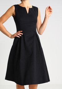 Robe noire sans manches avec un décolleté en V, corsage ajusté et jupe évasée. Tissu texturé avec de subtils motifs, design à hauteur des genoux.