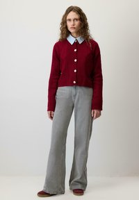 Czerwony sweter z dzianiny z wzorem w warkocze, zapinany na guziki, nałożony na jasnoniebieską koszulę z kołnierzykiem. Szare spodnie o szerokiej nogawce i bordowe buty.