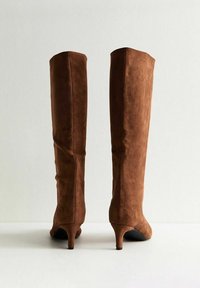 Stivali alti in suede marrone con punta appuntita e tacco moderato. La superficie presenta una consistenza morbida e dettagli di cucitura minimi.