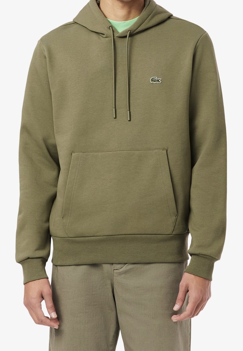 Homme portant un sweat à capuche vert olive avec poche avant et petit logo crocodile sur la poitrine, assorti à un pantalon vert.