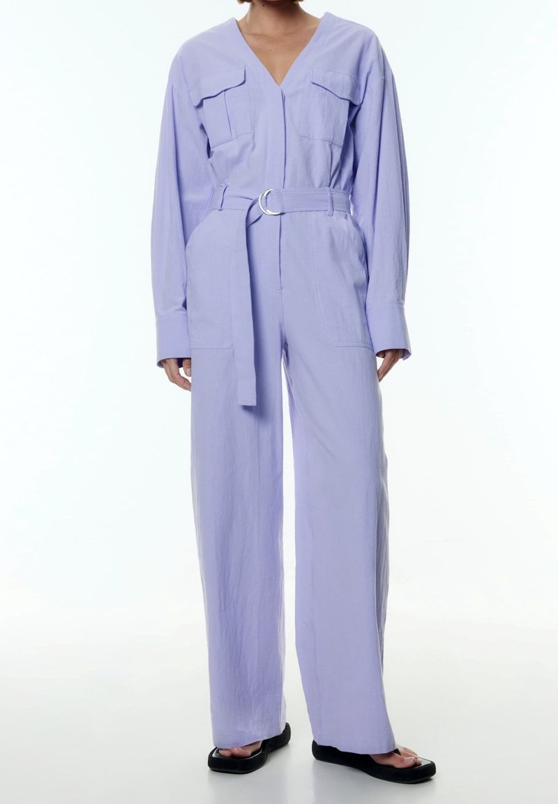 Femme portant une combinaison ceinturée violet clair avec manches longues, col en V, poches avant et sandales noires à bout carré.