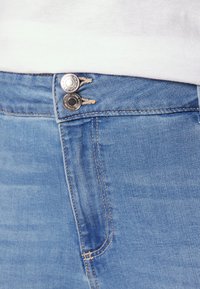 Jeans din denim albastru deschis, cu fermoar și două butoane metalice. Finalizate cu cusături contrastante și cu un buzunar pe lateral.