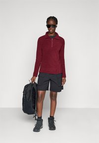 Bordeaux fleece pullover met een half rits, gecombineerd met zwarte cargoshorts en stevige zwarte wandelschoenen, terwijl hij een zwarte rugzak vasthoudt.