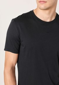 Crna majica kratkih rukava od glatkog pamučnog materijala. Okrugli izrez s prilagođenim krojem, prikazuje rame i dio ruke.