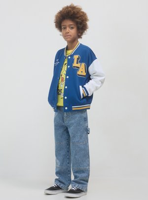 Giacca varsity blu con maniche bianche, lettering "LA" giallo, abbinata a una maglietta gialla e jeans baggy in denim azzurro chiaro. Sneakers nere.