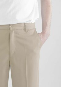 Beige Hose mit glatter Textur, ausgestattet mit einem Knopfverschluss, Seitentasche und einem maßgeschneiderten Schnitt, getragen mit einem weißen Hemd.