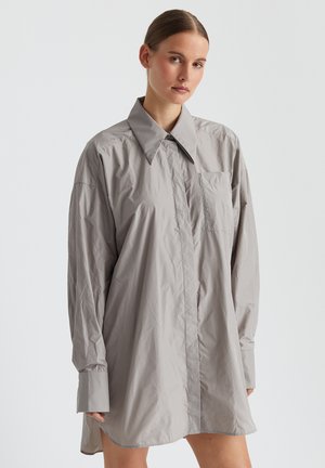 Hellgraues, oversized Hemdkleid mit spitzem Kragen, langen Ärmeln, vorderem Knopfverschluss und einer einzigen Brusttasche. Glatte Textur.