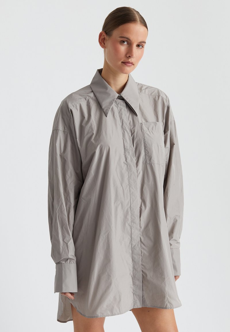 Hellgraues, oversized Hemdkleid mit spitzem Kragen, langen Ärmeln, vorderem Knopfverschluss und einer einzigen Brusttasche. Glatte Textur.