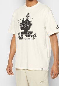 Bílá bavlněná tričko s černým grafickým designem stylizovaných květin a postav s textem "Bloom and ZOOM". Krátké rukávy a volný střih.